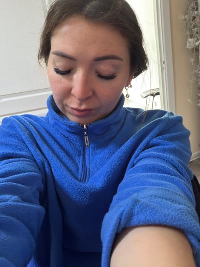 This is a close-up, waist-up, selfie-style portrait of a woman wearing a blue fleece quarter-zip jacket. She is looking down with her eyes closed and has on false eyelashes and dangling earrings. The background is out of focus, but appears to be indoors, possibly in a residential setting. There are no identifiable landmarks or features that place the image in or around the city of Япрыково, Russia.
