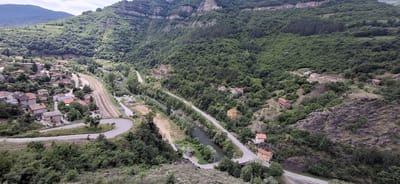 The image depicts a view of the Iskar Gorge near the city of Bov, Bulgaria. The scene is dominated by steep, forested hillsides, with a town situated on the flatter ground to the left. A railway line runs alongside the town and along the Iskar River, which can be seen winding through the valley. A road also follows the river's course. Several buildings are scattered along the hillsides, including residential houses and possibly some commercial structures. The foreground shows a winding road. The weather appears to be overcast, with a cloudy sky visible in the background.