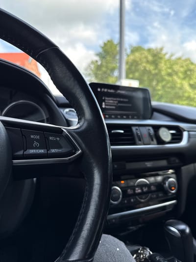 The image shows the interior of a car, focusing on the steering wheel and dashboard. A person's leg, wearing jeans with white speckles, is visible in the lower right corner. The steering wheel is black leather and has control buttons on the left side, including "MODE," "RES/+," "OFF/CAN," and "SET/-". A car's infotainment screen is visible in the center, displaying text that appears to be related to audio playback. The air vents and climate control buttons are also visible on the dashboard. Outside the car, green trees and a blue sky with clouds are visible through the windshield. The image does not provide any specific clues to pinpoint its location in Šilutė, Lithuania.