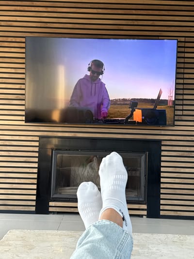 The image shows a person's legs in white socks and blue jeans, resting on a light-colored tiled floor. The feet are positioned in front of a black-framed fireplace, with a view of the flames inside. Above the fireplace is a flat screen TV displaying a DJ mixing music on a stage outdoors, with a bright blue sky in the background. The wall is covered in horizontal strips of wood with black trim. The scene is located in Daliyat al-Karmel, Israel, at coordinates 32.689, 35.054. The image is taken from a low angle, looking up at the person's legs. The environment is warm and inviting, suggesting a comfortable and relaxed atmosphere.