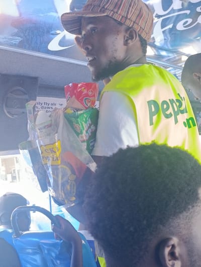 Inside a public transport vehicle in Matungulu west ward, Kenya, a male vendor stands, illuminated by bright daylight from a window. He is a dark-skinned individual, appearing to be in his 20s or 30s, with a short beard and a multi-colored striped cap (brown, orange, light blue, white). He wears a white t-shirt underneath a bright neon yellow-green sleeveless vest featuring the green text "Pepa" on its back. His mouth is slightly open, indicating he may be speaking. He holds a large, clear plastic bag containing various snack packets, including visible brands "KALUMA CHIPS" and "PICKS," along with other red and green-packaged items. In the lower right foreground, the back of a second individual's head with short, dark, curly hair is visible. On the far left, a third individual, wearing a black cap and light blue attire, is partially visible, appearing to be seated with hands on a blue, curved object, possibly part of a child's seat or toy. A fourth individual's head is partially visible behind the vendor's right shoulder. The background through the window includes a partially visible blue and white advertisement.