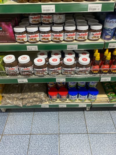 This image captures an aisle within a retail store, likely a supermarket or general store, in Goderich, Sierra Leone. The scene is illuminated by artificial indoor lighting, with no people visible, focusing entirely on the displayed merchandise.
The shelves are well-stocked with various packaged food items. On the upper left, a blue container of "Horlicks" malted milk drink is visible. Below it, numerous jars of "Nutella" hazelnut spread, distinguished by their white lids and familiar branding, are neatly arranged in multiple rows. Adjacent to these, on a parallel shelf, are rows of bottles, some labeled "RIYA" and "ZIYA," possibly containing honey or syrup. Further down this shelf, more "Nutella" jars are displayed.
Below the "RIYA" and "ZIYA" bottles, a selection of cans with distinct blue and red lids is stacked, some clearly marked with the brand "Estha," suggesting canned goods like fish or other food items. On the bottom shelf, large transparent plastic bags filled with what appears to be dried foodstuffs or bulk snacks are stacked. A partial view of a box at the bottom left reveals text that includes "the finest ted on earth."
The floor on the right side of the aisle is covered with light blue, speckled tiles. Green shelf dividers segment the product displays. Several white price tags are attached to the shelves, displaying numbers such as "245," "199," "275," "105," "110," "115," and "120," indicating the pricing of the goods. The overall arrangement suggests a typical, organized retail environment where products are presented for sale to customers.