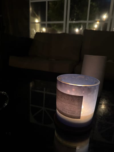 The image depicts an indoor scene with a lit candle in the foreground. The candle is in a glass jar and has a label that reads "LAND + FOG ON THE CALIFORNIA COAST". Behind the candle, a blurred background shows a couch and a window with lights visible outside, suggesting it is nighttime. The surface the candle rests on appears to be a dark, reflective table. There is no discernible location in the city of Colchester, United Kingdom within this image.