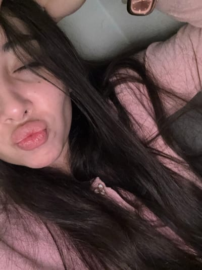 A person's face is visible with puckered lips, nestled amongst dark hair. The skin appears fair, with minimal makeup evident. They are wearing a light pink garment that is decorated with floral details. There is what seems to be a wristwatch in the image on the left side near the edge of the picture.