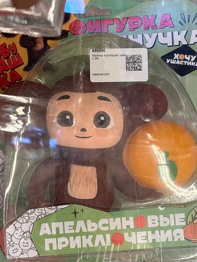 The image shows a close-up of a toy package featuring a Cheburashka character holding an orange. The toy is enclosed in clear plastic packaging. The packaging displays text in Russian, including "ФИГУРКА ЧЕБУРАШКА" (Figurka Cheburashka) and "АПЕЛЬСИНОВЫЕ ПРИКЛЮЧЕНИЯ" (Apel'sinovye Priklyucheniya - Orange Adventures). A label on the packaging has the number "446994" and a QR code. The background appears to be a retail environment. The image does not contain any information that allows for the determination of its location within the city of Kuzmolovsky, Russia.