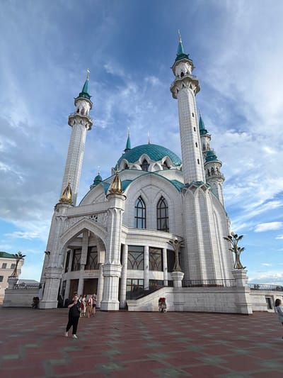 This is a photograph of the Kul Sharif Mosque in Kazan, Russia, taken on a bright, partly cloudy day. The mosque, a prominent landmark, is depicted from a low angle, emphasizing its grand architecture. Two tall, slender minarets with teal-colored spires flank the main building, which features ornate white stonework and turquoise domes. The exterior walls are adorned with intricate carvings and arched windows.
In the foreground, a red brick plaza extends from the mosque. On the plaza, a woman wearing a black top and pants is standing and looking at her phone. Behind her, near the entrance of the mosque, a group of people are gathered. The sky above is a vibrant blue, with wispy white clouds scattered across it, suggesting it's midday. The overall scene conveys a sense of architectural grandeur and a peaceful atmosphere. There is no visible text in the image.
