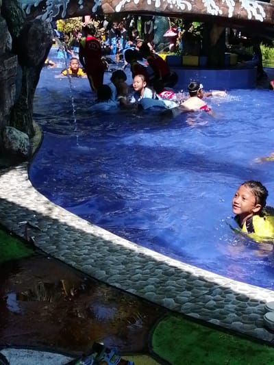 This is a photograph taken at a water park, possibly located in Tegalwangi, Indonesia, showing several children and adults swimming in a bright blue pool. In the foreground on the right, a young boy wearing a yellow life vest is smiling and looking upwards while swimming. To his left and further back in the pool, another child in a light blue shirt is partially submerged, with a red float visible. Several other individuals, including adults in red and blue attire, are also present in the water, some appearing to be supervising. The pool has a stone-like edge on the left side, and beyond the water, there is lush green grass. The background includes decorative elements resembling rocks and trees, suggesting a recreational setting. The water is rippling, indicating movement.