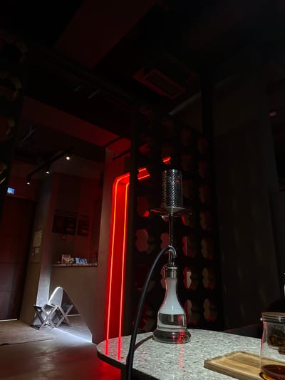 The image depicts the interior of a dimly lit establishment, likely a hookah lounge or cafe, with red ambient lighting. The foreground features a granite-patterned table with a hookah. The hookah has a clear glass base filled with water, a metallic stem, and a perforated metallic bowl or heat management device on top. A black hose extends from the base. To the right of the hookah, on the same table, is a glass vessel, possibly a teapot or another serving dish, with a wooden lid and some contents visible inside.
In the background, to the left, is a counter area with items on it, suggesting a service point. Further left, there is a closed door with a blue sign reading "ВХОД" (Entrance). Adjacent to the door is a tall, grey architectural element that frames an entrance or alcove, within which are two folded dark chairs. A bright vertical red neon light runs along the side of this architectural element. The walls are decorated with a repeating pattern of cutouts or illuminated shapes. The ceiling is dark, with some visible structural elements and lighting fixtures. The overall atmosphere is moody and atmospheric, typical of an evening entertainment venue. The location is identified as Бородино, Russia.