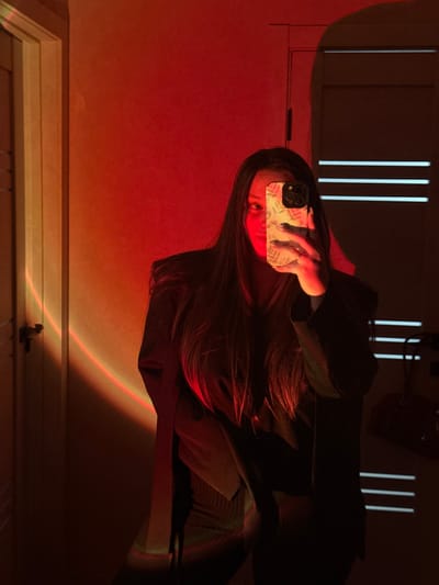 An indoor scene in Tliustenkhabl', Russia, depicts a person taking a mirror selfie. The subject, a female with long dark hair, wears a dark top and an oversized dark blazer. She holds a smartphone in her right hand, obscuring most of her face except for her left eye and forehead. The smartphone features a light-colored case with a floral or leafy pattern and a visible triple-lens camera module. A ring is present on her right ring finger. The environment is illuminated by a projected light source, creating a prominent red and orange circular gradient on the wall to the left of the subject. A white door frame is visible on the far left. To the right of the subject, a dark vertical surface, possibly another door or a panel, exhibits three horizontal light-colored stripes. The overall lighting conditions are dim, with the projected red light dominating the scene.