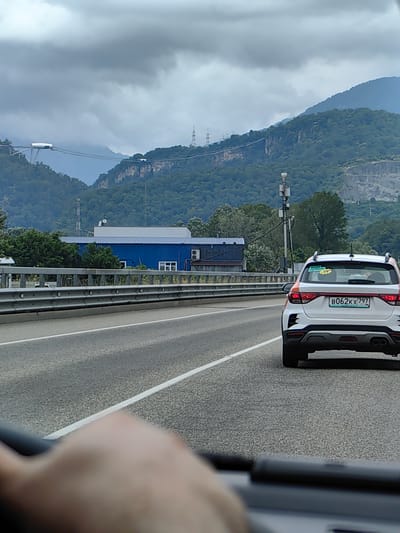 The image shows a view from a vehicle on a highway with a white car in front. The white car has a Russian license plate that reads "B062KX 797". In the background, there are lush green mountains, a blue building, and power lines. The sky is cloudy and overcast. This scene appears to be in the city of Vysokoye, Russia, given the context of the license plate and the mountainous terrain.