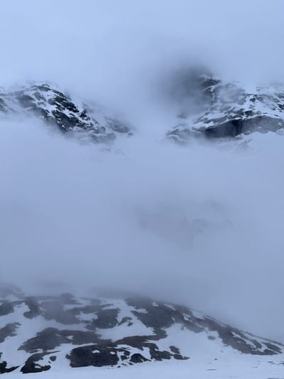 The image presents a dramatic alpine landscape, featuring two prominent, rugged mountain formations partially obscured by dense, light grey fog or low-hanging clouds. The mountains, visible on the left and right sides of the frame, exhibit stark contrasts between dark, exposed rock and irregular patches of white snow, indicating steep and complex terrain. The texture of the snow and rock is detailed, showcasing the harsh environment.

The central and larger portion of the image is enveloped in a thick, ethereal mist, which diffuses the light and creates a sense of depth and mystery. Tendrils of fog also creep along the edges of the mountain peaks, softening their harsh outlines. The overall lighting is soft and uniform, suggesting an overcast day. The presence of significant snow cover points to cold weather conditions, likely winter or early spring/late autumn.

The scene captures a feeling of vastness and isolation, emphasizing the untamed beauty of nature. No people, animals, or man-made objects are visible, reinforcing the remote and pristine character of the location. Given the context, this is a typical high-altitude mountain scene in Poschiavo, Switzerland. No text is visible in the media file.