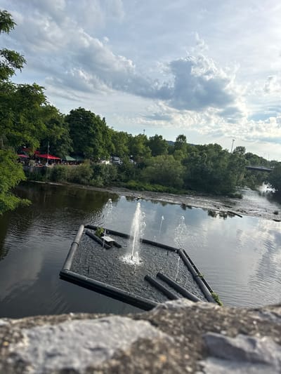 The image depicts a wide-angle view of the Lahn river in Wetzlar, Germany, as seen from a stone bridge or embankment. In the foreground, out of focus, is a textured grey stone surface. The river occupies the central and lower portions of the image. In the middle of the river, a rectangular structure made of black pipes supports a fountain that is actively spraying water upwards. Smaller fountains are also visible around the perimeter of the structure.

Along the left bank of the river, dense green trees line the water's edge. Below the trees, a section of a waterfront area is visible with several people seated at tables under red and green umbrellas. Further back, parked cars are visible.

The right side of the river features a more natural, less developed bank with trees and scrub. In the distance, a bridge spans the river, with traffic visible on it. The sky above is partly cloudy, with streaks of white clouds against a pale blue background. The overall impression is that of a serene, possibly recreational, outdoor setting.