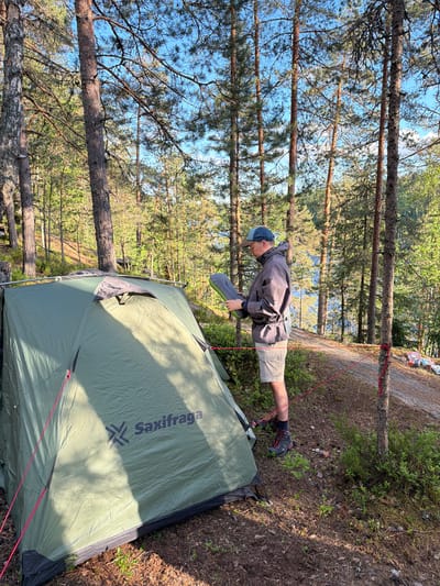 A person wearing a cap, jacket, shorts, and boots stands next to a green tent in a forest. The person is holding a deflated sleeping pad. The tent has the brand name "Saxifraga" visible. The ground is covered with pine needles and small plants. In the background, there are tall pine trees and a body of water. The scene appears to be a campsite. The location is Berezovo, Russia.