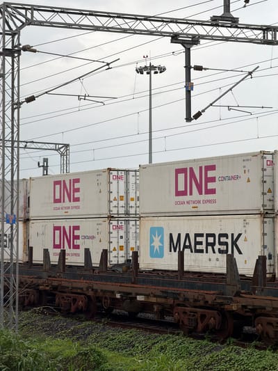 The image captures a freight train loaded with shipping containers, passing through Saphale, India. The train features multiple stacked containers. The top row of containers displays the "ONE OCEAN NETWORK EXPRESS" logo in large pink lettering. Below these, a container with the "MAERSK" logo is visible. The train cars are old and rusty. Overhead, a complex network of electrical wires and support structures for an overhead catenary system is present, indicating the railway is electrified. The sky is overcast. The surrounding area appears to be a railway embankment with sparse green vegetation.