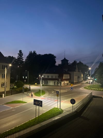 The image depicts a street scene in Ostróda, Poland, during twilight. A four-way intersection is visible, with a roundabout in the center. Several buildings, a pedestrian crossing, streetlights, and traffic signs are present. Two individuals are walking on the sidewalk, and a car is visible on the road. The sky is a deep blue, indicating the transition from day to night, and streetlights cast a warm glow on the surroundings. The overall atmosphere is calm and serene.