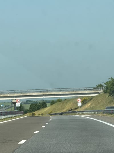 The image displays a section of an asphalt road, likely a highway, in Vratitsa, Bulgaria, under a clear, pale blue sky. A concrete overpass with metal railings traverses the frame horizontally in the mid-ground. The road surface exhibits white dashed and solid lane markings, delineating a main thoroughfare and a curving exit or entry ramp on the left. Metal guardrails are present along both sides of the road. Beneath a structural support of the overpass on the left, a circular regulatory sign with a red border and black truck icon indicates a restriction against trucks, supplemented by a rectangular sign displaying "1500m" and "7h-19h". An identical truck restriction sign, also accompanied by a rectangular sign partially visible with "7h-19h", stands on the right grassy embankment. The environment consists of green, grassy slopes rising on either side of the road, interspersed with trees and bushes. A faint white vehicle is visible in the far distance on the left side of the road. No individuals or active vehicles are present in the foreground or mid-ground.