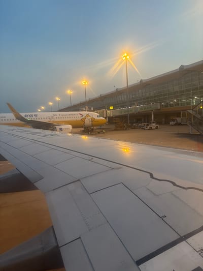 An aerial view of an airport tarmac at dusk, featuring a passenger airplane with yellow and white livery, a modern airport terminal building with large glass windows, and several airport vehicles. The airplane's tail is visible on the right side, with the text "Air Travel 湖南航空" and an Airbus A320 model number. Yellow lights illuminate the tarmac and the building. The foreground shows the wing of another airplane, a portion of which is in focus. The sky above is a soft blue with hints of orange from the setting sun, and the sun's rays are visible, creating a lens flare effect. The scene is likely at an airport in Xinwu District, China, given the context provided by the text on the airplane.