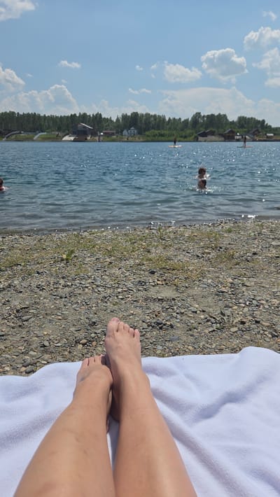 The image depicts a scene at a lake in Verkh-Katunskoye, Russia. In the foreground, a pair of legs and feet are resting on a white towel on a pebbly shore. The lake is calm with ripples on the surface and reflects the bright blue sky with scattered white clouds. In the middle ground, several people are visible in the water, some swimming and others stand-up paddleboarding. Along the far bank of the lake, there is a line of trees and several small buildings, suggesting a recreational area or a small settlement. The overall impression is of a sunny day at a lake with people enjoying outdoor activities.