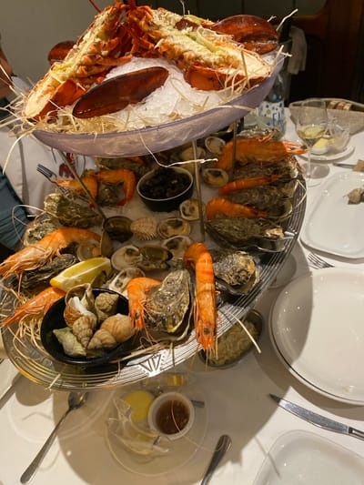 The image displays a multi-tiered seafood platter, a "fruits de mer," centrally positioned on a white-clothed table within a restaurant setting in La Wantzenau, France. The uppermost tier features two split, cooked lobsters arranged on crushed ice, bordered by natural straw. The middle tier presents a selection of cooked prawns, oysters, and clams, alongside a small black bowl containing small, dark-shelled mollusks. The lowest tier holds additional oysters, prawns, and a black bowl filled with various sea snails, including whelks, accompanied by lemon wedges.
The table environment includes several white ceramic plates, some empty, some with residual food. Silverware, consisting of forks, knives, and spoons, is present. A wine glass containing white wine and a water bottle labeled "EAU" are visible. Small condiment dishes containing lemon slices and a brown dipping sauce are also on the table. The upper left quadrant of the image shows the shoulder and arm of an individual, clad in a white shirt.