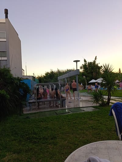 The image depicts an outdoor scene at a resort in Termas del Daymán, Uruguay. In the foreground, there is a green lawn, with the corner of a white plastic table visible. In the middle ground, a group of people are using a multi-shower facility. Some are sitting on a bench under the showerheads, while others are standing nearby. To the right, there are more people near a pool area with white umbrellas and green lounge chairs. A palm tree stands between the showers and the pool. In the background, there is a building on the left with a chimney and several windows. Lush trees and shrubs are also visible. The sky is a gradient of blue and orange, indicating either dawn or dusk.
