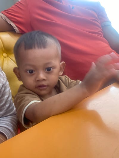 A young child with short, dark hair, wearing a brown shirt, sits at an orange table. The child is looking towards the camera, with their right hand blurred due to motion. In the background, a person in a red shirt is partially visible, seated on a yellow chair. The scene appears to be indoors, possibly a restaurant or home setting. There is no identifiable information about the specific location within Kudus, Indonesia.