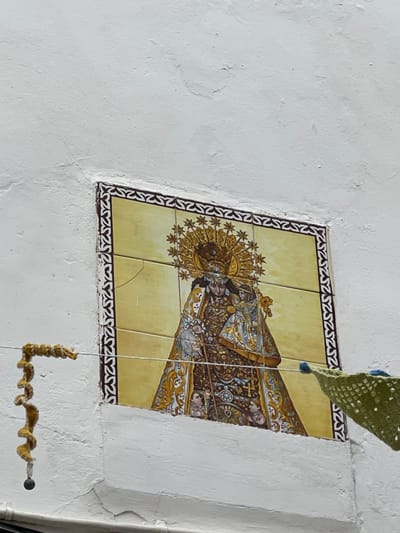 The image captures a weathered white stucco building wall in Buñol, Spain, featuring a rectangular ceramic tile panel embedded in its surface. The panel depicts a crowned female figure, likely the Virgin Mary, holding a smaller crowned figure, presumably the infant Jesus. Both figures are adorned in richly detailed robes with gold, brown, white, and blue accents. A prominent golden sunburst halo with radiating points encircles the main figure's head, set against a light yellow background. The tile panel is framed by a repeating dark brown patterned border. Multiple vertical and horizontal cracks are visible across several tiles within the panel. Two thin horizontal wires, likely clotheslines, stretch across the lower portion of the scene in front of the tile panel. From the left wire hangs a coiled, yellow-beige knitted or crocheted ornament, terminating in a small dark sphere. A section of a green, loosely woven or crocheted fabric item, possibly a decorative flag or garment, hangs from the right wire, partially visible on the right edge of the frame. The white wall exhibits minor cracks and texture variations, indicating its age and exposure. No individuals are present in the scene.