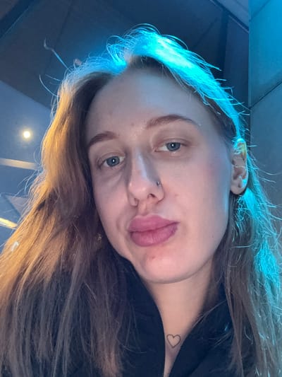 A close-up, angled selfie photograph depicts a fair-skinned young woman. She has light brown hair with blonde highlights and blue eyes. She has a nose piercing on the left side of her nose. A small, heart-shaped tattoo is visible on the right side of her neck. The lighting in the scene appears to be predominantly blue. The background is blurred and indistinct. There is no evidence of the location being Ol'ginskaya, Russia.