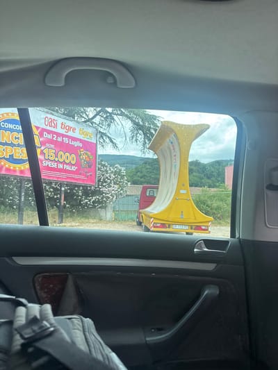 The image, taken from inside a vehicle in Ascoli Piceno, Italy, provides a view through the rear window. The dominant subjects are a large, yellow, curved amusement park ride component, possibly a part of a roller coaster or a similar attraction, and a billboard advertising a lottery or promotion.
The yellow ride component is prominently displayed, with its distinctive shape dominating the upper portion of the outdoor scene. It appears to be a structural element, possibly made of fiberglass or a similar material, with some water droplets visible on its surface, suggesting recent rain or dew. Behind this, a red truck is partially visible.
The billboard is advertising "Oasi tigre tigre," with the dates "Dal 2 al 15 Luglio" (From July 2nd to July 15th). It highlights a prize of "15.000 SPESE IN PALIO*" (15,000 expenses in play*). The text "AMICO" is also present. The billboard's design includes imagery of a shopping cart filled with products, reinforcing the idea of a prize related to spending or shopping.
The setting is outdoors, with lush green hills and trees in the background, under a cloudy sky, suggesting it's daytime. The overall scene suggests a fairground, amusement park, or a promotional event taking place in a rural or semi-rural Italian landscape. The interior of the car, with its door handle and grey trim, frames the outdoor view. The image is oriented vertically.