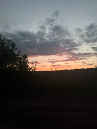 The image shows a sky with a dramatic contrast between a dark, overcast area with low-hanging clouds on the left and a sunset with orange and pink hues on the right. Thin power lines stretch across the cloudy portion. A line of trees is silhouetted against the sunset. A dark, indistinct foreground occupies the right side and bottom of the image. The setting appears to be rural due to the presence of trees and open sky.
I don't have the location information for Галево, Russia, I don't have any details about any people.
