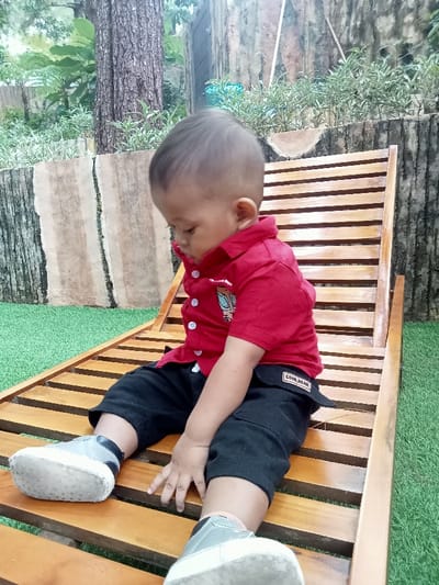 An infant is seated on a wooden slatted lounge chair in an outdoor garden setting. The scene is consistent with a private or public recreational area in Kuwukan, Indonesia.

The subject is a single infant, appearing to be male, dressed in a red short-sleeved collared shirt with white buttons and a light-colored graphic on the left chest, black cargo shorts with a "COOL BEAR" label on the left pocket flap, and grey and black baby shoes with white soles. The infant's head is tilted downward, directing its gaze towards its lap or the chair. Both hands rest on its thighs.

The immediate ground surface is covered with green artificial turf. Behind the infant and chair, a low, textured wall, resembling aged wood or concrete, serves as a planter border, containing small green plants with white variegation. To the left, a portion of a large tree trunk with rough bark is visible. Further in the background, additional green foliage and trees indicate a natural or landscaped environment. Natural daylight illuminates the scene. No other individuals or active interactions are observed.