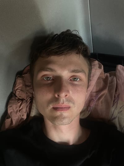 The image is a close-up, rotated portrait of a young man lying down, possibly in bed, with his head resting on a pillow. The background includes a textured grey surface to the left and a patterned fabric, likely a duvet or blanket, in shades of pink and beige, to the right. The man has light skin and light brown hair. His eyes are open and directed towards the camera. He has a light stubble on his face and his lips are slightly parted. The lighting appears to be artificial, suggesting an indoor setting, possibly at night or in a dimly lit room. No text is visible in the image. The man's expression could be interpreted as neutral or perhaps tired. The overall scene is intimate and personal.