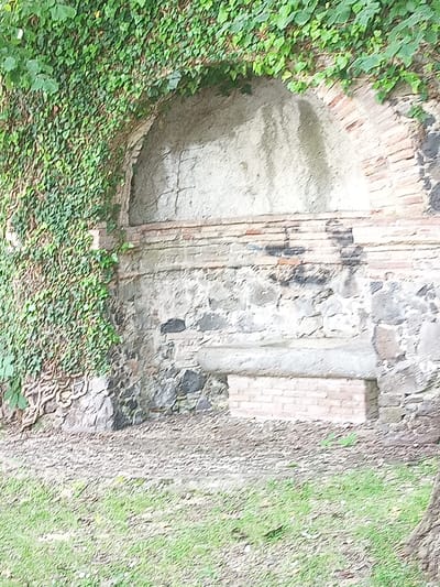 The image shows a stone structure partially covered in ivy, with an arched opening. The opening appears to be a niche or alcove, possibly part of a larger ruin or garden feature. The stonework is varied, with some sections appearing to be rough-hewn stone and others made of brick. A flat ledge or shelf is visible within the lower part of the niche, and a smaller brick structure is located in front of it. The ground in front of the structure is covered with a mixture of soil, gravel, and grass. The overall impression is of an old, weathered structure in a natural setting. The context of the city of Olot, Spain, suggests this might be a historical garden or park feature, possibly related to the volcanic landscapes for which the region is known. However, without further information, specific details about its purpose or origin within Olot cannot be determined from the image alone.