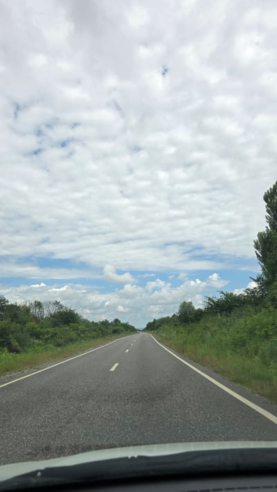 The image depicts a view from a vehicle traveling on a paved road through a rural area. The road has a dashed white line in the center and a solid white line along the right edge. Lush green vegetation lines both sides of the road. In the distance, a lone person appears to be walking on the side of the road. The sky is filled with white, fluffy clouds against a blue sky. The scene is consistent with a road in the vicinity of Goyty, Russia, a region characterized by its natural landscapes.