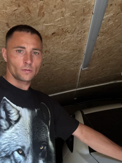 A man with short, dark hair and green eyes is shown in a selfie. He is wearing a black t-shirt with a large wolf graphic. His left arm is extended, resting on a light-colored car. The background consists of a textured, wooden ceiling with a fluorescent light fixture. The man appears to be inside a garage or workshop.