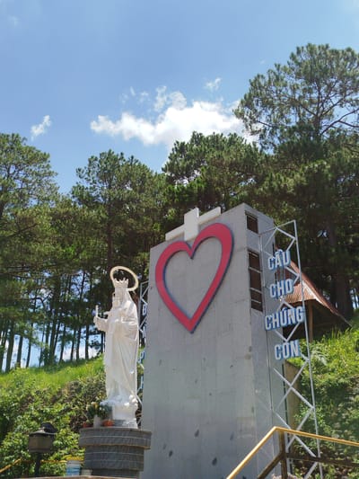 The image depicts a large outdoor statue of Mary holding the infant Jesus, standing on a circular, multi-tiered base. The statue is white and detailed, with Mary wearing a crown and a halo. Behind the statue is a large, grey, rectangular monument featuring a large red heart outline with a white cross at the top. To the right of the monument, blue text on white poles reads "CẦU CHO CHÚNG CON." The scene is set against a backdrop of lush green trees, likely pine trees, under a bright blue sky with scattered white clouds. The ground is covered in grass and foliage, and a metal railing is visible in the foreground. The overall atmosphere is serene and religious, suggesting a place of worship or a pilgrimage site. The text on the monument translates to "Pray for us," a common Catholic supplication. This monument is likely located in K'Rèn, Vietnam, a region known for its religious sites and natural beauty.