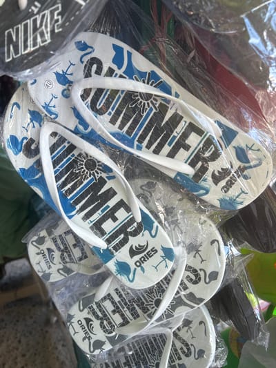 The image displays a close-up of several pairs of flip-flops packaged in plastic, likely at a market stall in Bira, Indonesia. The flip-flops have white soles with blue and black designs. Some soles feature the word "SUMMER" in large, black lettering with a sun graphic above it. Other soles have the brand name "ORIES" and graphics of flamingos and other stylized animals. A black cap with the "NIKE" logo is visible at the top left of the frame. The overall setting appears to be a retail environment, with the items being displayed for sale.