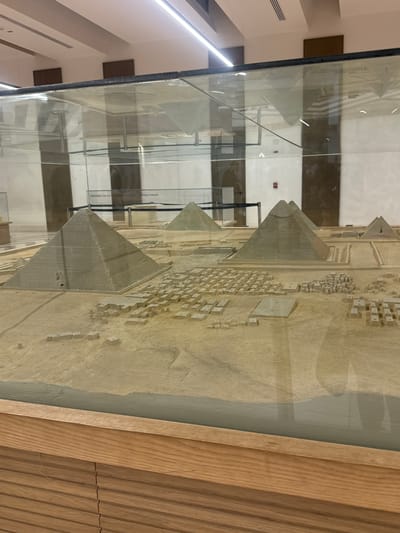 A detailed model of the pyramids of Giza is enclosed in a glass display case with a light-colored wooden frame. The model includes the pyramids and surrounding buildings, possibly representing the layout of the city of Giza, Egypt. The glass case is in a modern-looking building with light-colored walls.