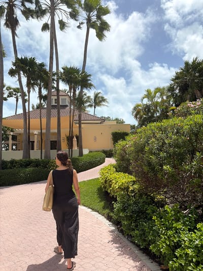 A medium, eye-level shot, rotated to the left, showcases a woman walking along a red brick path lined with green vegetation and palm trees, with buildings visible behind the trees. The sky is blue with scattered white clouds.
The woman, dressed in a black dress and sandals, is walking away from the camera, carrying a light-colored bag. She has curly brown hair.
To the left of the path, tall palm trees line the road, obscuring parts of the buildings behind them. The buildings are painted in light yellow and beige tones, with tiled roofs and visible windows.
The path itself is made of red bricks in a herringbone pattern. The green vegetation, consisting of bushes and trees, lines the path on both sides. The image appears to be taken in Key Biscayne, United States of America.