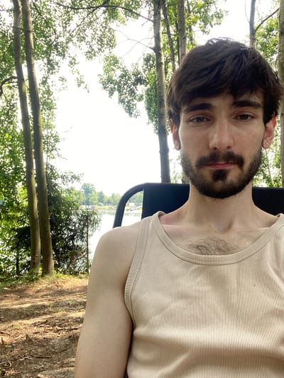 A young adult male with dark hair and a beard is sitting outdoors in Kaniów, Poland, by a body of water. He is wearing a light-colored ribbed tank top and appears to be looking towards the camera. The background consists of trees, foliage, and a lake. The lighting suggests it is daytime.