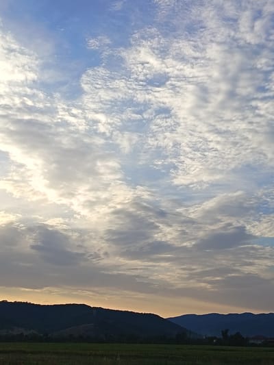 This is an eye-level, full shot of a landscape at Skravena, Bulgaria, under a dramatic sky with scattered clouds. The foreground features a dark green field, possibly with crops, bordered by rolling hills and mountains. The horizon is marked by a band of warm, golden light from the setting sun, which is peeking through the clouds. The sky is a gradient of blues, with lighter shades near the horizon and darker blues towards the top, filled with a variety of white and gray clouds, some of which are wispy and scattered, while others are more dense and textured. The overall impression is one of natural beauty and tranquility.