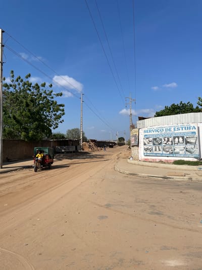 The image displays a sunny daytime scene on an unpaved dirt road in Luanda, Angola. The sky is a vibrant blue with a few wispy white clouds. Power lines and utility poles run diagonally across the frame, extending into the distance.
Along the left side of the road, a building with a white facade features a large sign. Further down, piles of dirt or debris are visible, and trees line the horizon. A green motor-tricycle, resembling a tuk-tuk, is positioned on the left side of the road, with at least one person wearing a yellow garment visible inside it. Another small, indistinct figure appears to be walking further down the road. The road surface is dry and dusty, showing visible tire tracks.
The time of day is clearly bright daylight, indicated by the intense blue sky and crisp shadows. The weather is clear and sunny.
The text visible on the building's sign, as per OCR, reads: "TAAN BI COMERCIO E SERVIÇOS (SU), LDA," followed by "SERVIÇO DE ESTUFA." Additional text includes "THA FORMACA," likely "ARTIGOS EM NOVOS," and contact numbers: "944674973-TONY / 937.912.470." The sign also displays illustrative images of vehicle interior components such as seats, steering wheels, and headrests, suggesting the business offers automotive upholstery or repair services (possibly relating to vehicle heating/cooling or interior customization, given "ESTUFA" which can mean 'furnace' or 'greenhouse' but here likely refers to vehicle interior-related service).