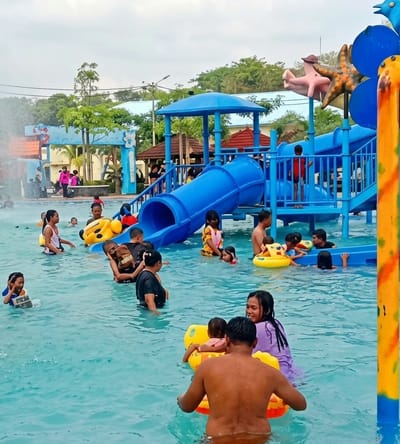 The photograph captures a vibrant scene at a water park in Durin Simbelang, Indonesia. The image is dominated by a blue water slide structure with a covered platform and decorative elements such as a pink figure wearing a hat, and blue starfish, attached to its top. Numerous people, including adults and children, are scattered throughout the pool. Some are swimming freely, while others are using yellow inflatable tubes. A man and woman are assisting two children in a yellow float. The water is a light blue color, and the sky above is overcast. In the background, there are additional structures and trees, indicating the park's broader setting.