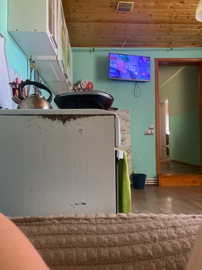 The image depicts an interior scene, likely a kitchen area, possibly located in the village of Afanasyevo, Russia, given the context provided by the user. In the foreground, a portion of a person's leg and a textured beige rug are visible, suggesting the viewer is seated or lying down.
The main focus of the image is a vintage, white stove. On top of the stove, a metallic kettle with a black handle is prominently placed. To the right of the kettle is a dark frying pan. Above the stove and extending to the left are a set of light-colored kitchen cabinets.
The wall behind the stove is painted a light teal color. Mounted on this wall, to the right of the cabinets, is a flat-screen television displaying a man's face. A black wire hangs loosely from the TV. Further to the right, a doorway with a wooden frame leads to another room, which appears to be a hallway or another part of the dwelling. The hallway is also painted in muted tones of green and beige, and the floor appears to be wooden.
In the lower right portion of the image, a dark green bucket is visible on the floor, next to a lime green towel hanging on the side of the stove. There are electrical outlets on the teal wall near the doorway. The ceiling is made of wooden planks. The overall impression is one of a lived-in, possibly older, domestic space.