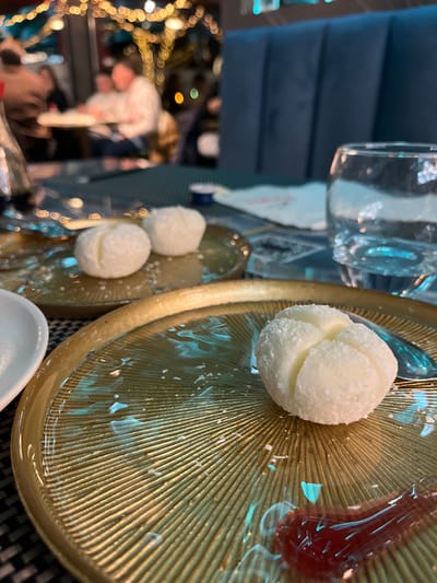 The image presents an indoor scene, likely a restaurant or café, with a focus on a dessert or snack presentation. In the foreground, two golden-colored, ribbed plates are visible. The larger plate, occupying the right side, holds a prominent white, spherical dessert. This treat appears to be a truffle or candy, covered in fine white flakes, possibly coconut, and features distinct indentations that give it a segmented or floral appearance. On the same plate, towards the back, there's a portion of a reddish, reflective substance, likely a fruit jelly or preserve.

A smaller, similar golden plate is positioned to the left, displaying two more of the white, spherical desserts, though less detailed than the one in the foreground. Scattered white flakes are visible across both plates. A small, white, ceramic spoon or dish is present in the bottom right corner. A clear glass is partially seen in the upper left, casting reflections.

The background is softly blurred, indicating a shallow depth of field. Visible elements include a dark blue or teal upholstered chair or booth seating in the upper left. Further in the background, several indistinct figures of people can be discerned, suggesting a populated dining environment. Numerous warm, blurry lights, appearing as bokeh, illuminate the left side of the scene, implying decorative string lights or ambient lighting, characteristic of an evening or night setting. No specific text is visible in the image. The overall atmosphere is warm and inviting, hinting at a pleasant dining experience in Basiglio, Italy.