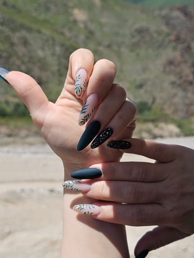 The image shows a pair of hands with elaborate nail art, positioned against a blurred background of hills and a sandy area. The hands are the primary subject, with the focus on the fingernails. The nails are long and almond-shaped, decorated in various styles. Some nails feature a pale pink or nude base with delicate green leaf designs and gold glitter accents. Others are painted in a matte dark teal or black, some with gold glitter sprinkled across them. One thumb nail appears to be a light blue with a hint of shimmer. The hands are positioned in a way that showcases the nail art, with one hand gently resting over the other. The background suggests an outdoor setting, possibly a beach or a dry, hilly landscape, but it is out of focus. There is no visible indication of the city of Tyrnyauz, Russia.