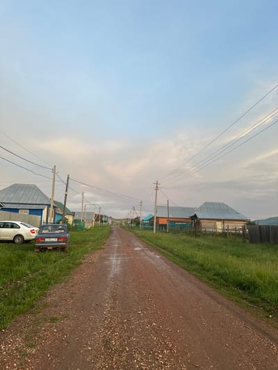 The image depicts a rural dirt road in what appears to be a village in Urazovo, Russia. The road is wet and muddy, suggesting recent rain. On the left side of the road, a white car and a dark blue car are parked. Both cars have Russian license plates. Beyond the cars, several houses with metal roofs are visible, some painted blue and others with natural wood or orange facades. A wooden fence runs along the side of the properties. Overhead, power lines stretch across the scene, supported by utility poles. The right side of the road is bordered by a grassy field with more houses and fences in the distance. The sky is partly cloudy with soft hues of pink and blue, indicating either dawn or dusk. The overall impression is one of a quiet, rural settlement under a transitional sky.