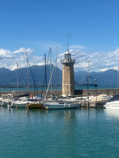 The image shows a harbor in Desenzano del Garda, Italy. In the foreground, there is a body of turquoise water with gentle ripples. Moored boats are visible along a stone quay in the middle ground. These boats include several white sailboats with tall masts, some with their sails furled. One sailboat stands out with a bright yellow hull. Behind the boats, a stone lighthouse with a lantern room at its peak is prominent. The lighthouse is surrounded by a stone wall. Beyond the quay and lighthouse, a body of water, likely Lake Garda, stretches towards distant mountains. The mountains are covered in vegetation and appear hazy. The sky above is a clear blue, dotted with fluffy white clouds, some of which are tinged with gray. The overall impression is a serene and sunny day in a picturesque Italian harbor.