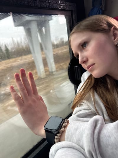 A blonde-haired woman with light skin is looking down in a photograph taken inside a vehicle. She is wearing a light grey hooded sweatshirt and an Apple Watch with a beige scrunchie around the wrist. Her hand is extended and resting on the windshield of the vehicle, which has water droplets on the glass. The reflection of a snowy urban environment can be seen in the windshield. The photo was taken inside the vehicle in Kukushtan, Russia.