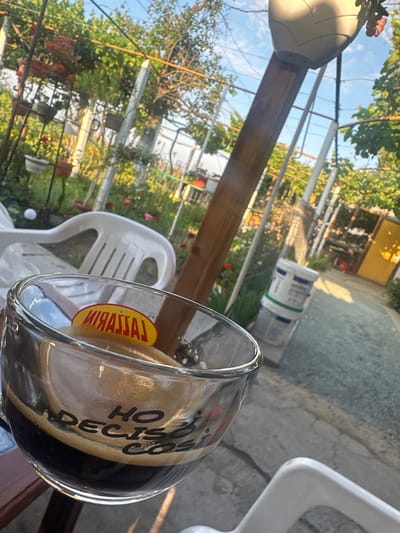 The image is a close-up, slightly tilted shot of a glass of dark liquid, likely coffee, with foam on top. The glass has writing on it, partially obscured by the drink. A yellow oval sticker with red lettering is visible on the inside rim of the glass. In the background, there is a garden area with greenery, flowers, and what appears to be a white plastic chair. A wooden pole with a large, light-colored object on top, possibly a lamp or a hanging planter, is prominent. A gravel path and some buckets are also visible. The scene suggests an outdoor setting, likely a patio or garden in Golyam Chardak, Bulgaria, given the context. The lighting indicates it is daytime, with sunlight filtering through the trees. There are no people visible in the image.