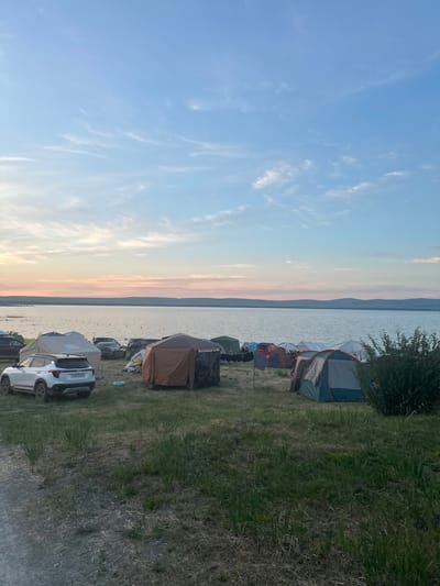 The image depicts a campground scene on the shore of a large body of water, likely a lake, under a sky with scattered clouds and the soft hues of either sunrise or sunset. Several tents are set up on the grassy terrain, varying in color and size, including a prominent tan and brown multi-room tent and a green and grey dome tent. Various vehicles, including a white Kia SUV and other cars, are parked among the tents. In the background, the calm water stretches to the horizon, with a distant, undulating shoreline. There are no visible people in the immediate foreground or mid-ground, though there might be figures in the water in the distance. The overall environment is that of an outdoor recreation area.